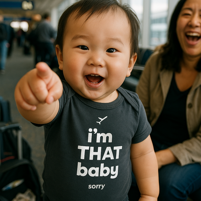 Baby wearing a onesie with text, standing in an airport with a smiling woman behind